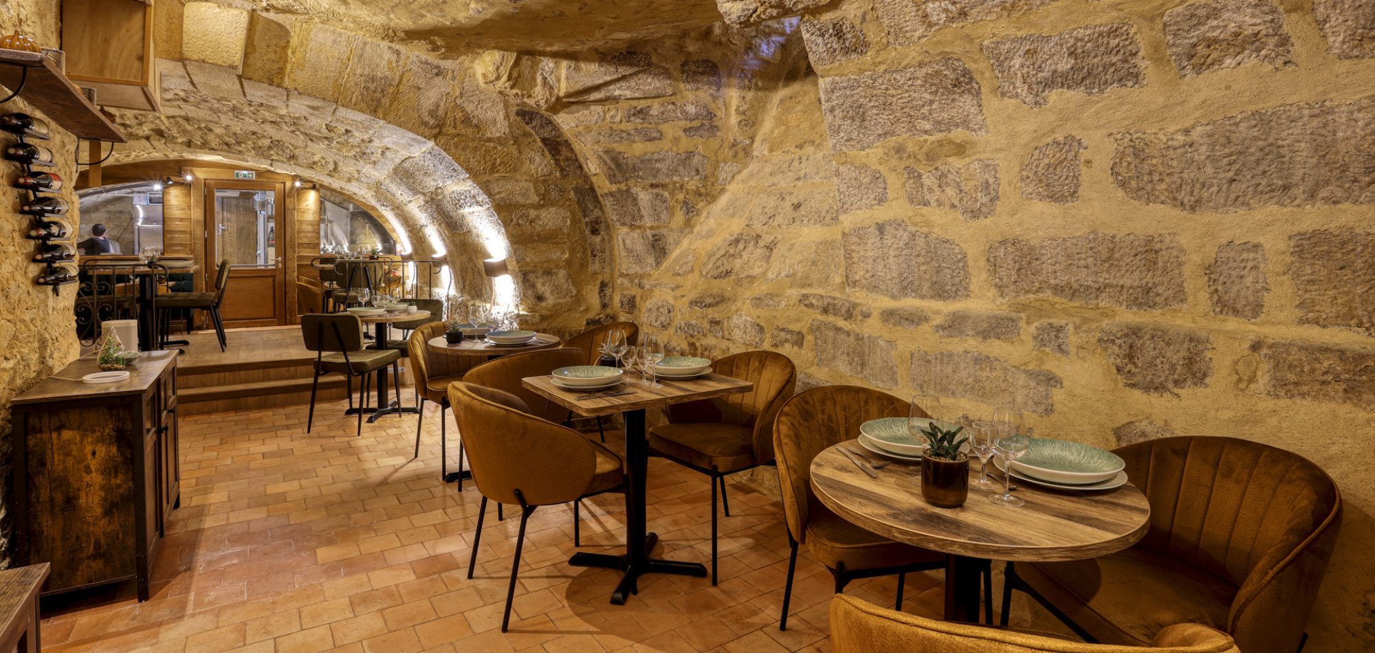 Salle voûtée du restaurant bar à vins Arcadia à Sarlat, fauteuils moutarde et tables dressées sous les pierres apparentes